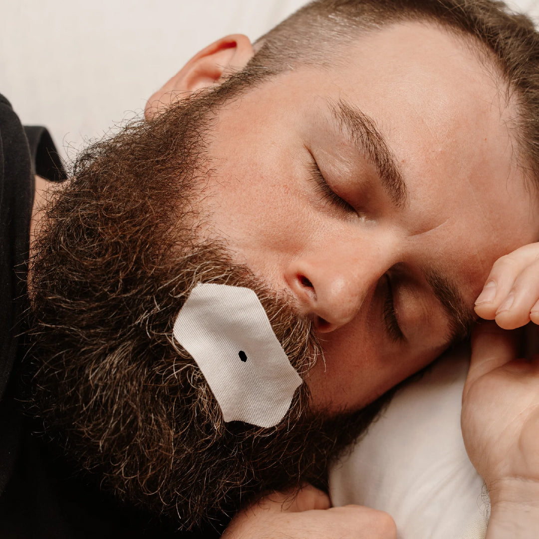 Man with a beard sleeping with Intake Breathing Technology LLC mouth tape applied to his lips.