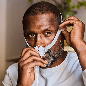 A close-up of a man wearing a white sleep apnea mask and adjusting the straps with both hands.