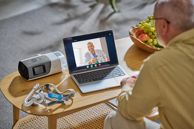 A man in a yellow shirt sits at a wooden table, using a laptop to video call a doctor. On the table next to him are a pulse oximeter and a mask
